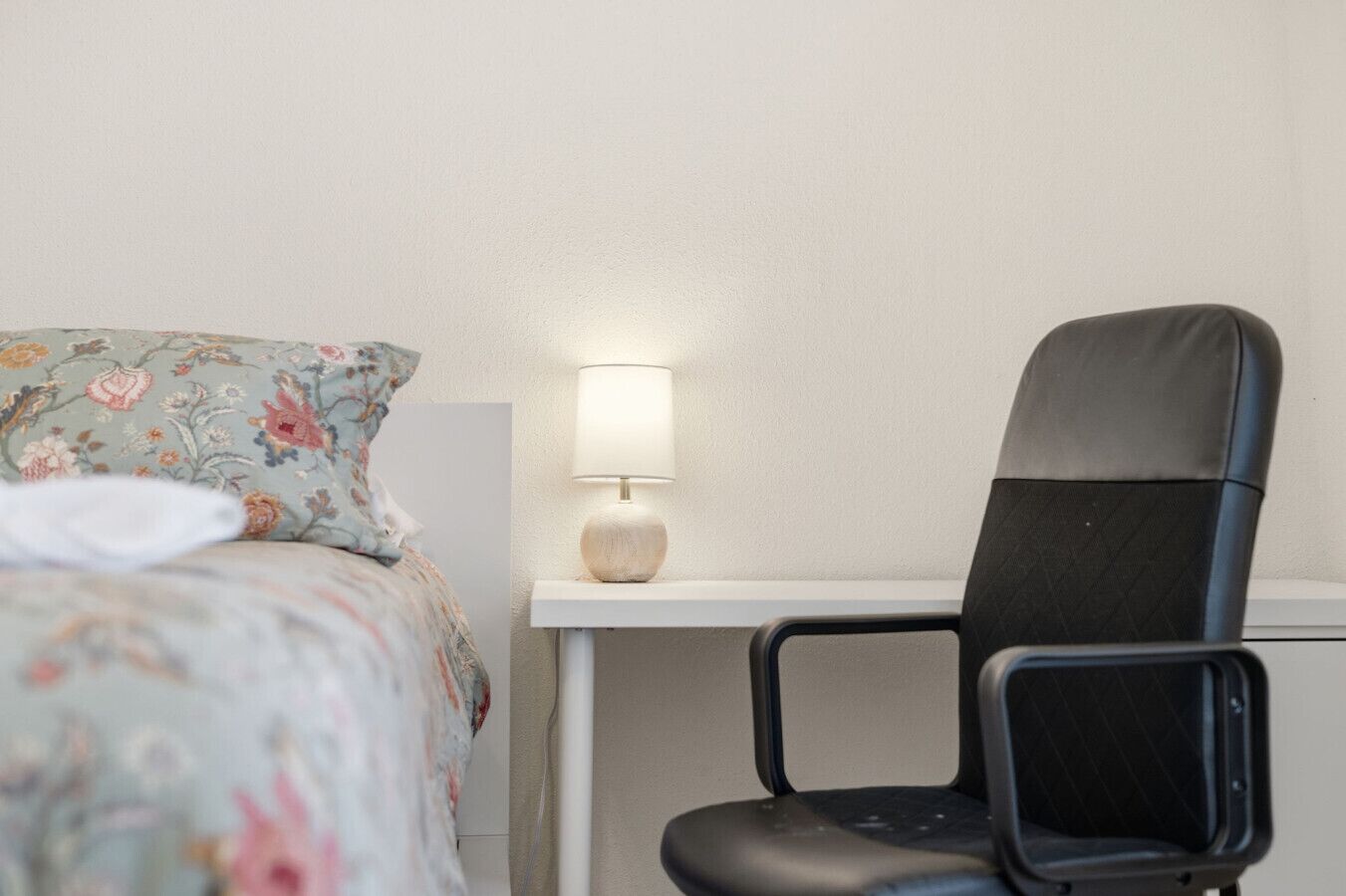A bed with floral bedding, a desk with a lamp, and a black office chair.