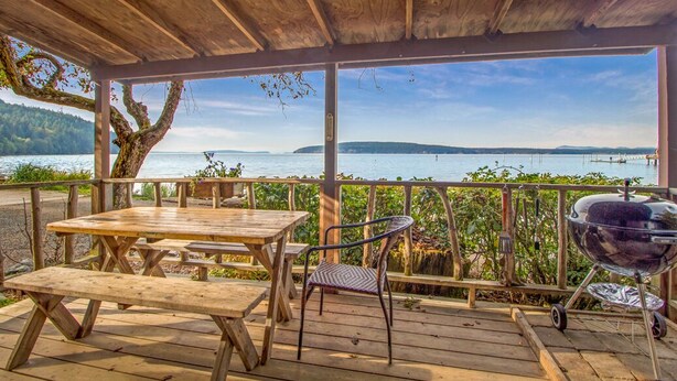 A wooden deck with picnic tables and a barbecue grill overlooking a lake. - 2BR Beachfront Cabin w/ loft - steps from prime west-facing beach! (Eastsound)
