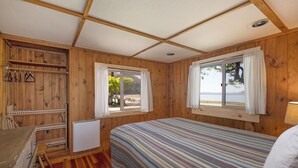 A wooden cabin room with a bed, a desk, and a chair. There are two windows with curtains, one of which offers a view of the beach. - 2BR Beachfront Cabin w/ loft - steps from prime west-facing beach! (Eastsound)