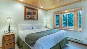 Un dormitorio con techo de madera, una cama grande con ropa de cama blanca, dos lámparas de noche, una ventana con vista a árboles y un cuadro en la pared.