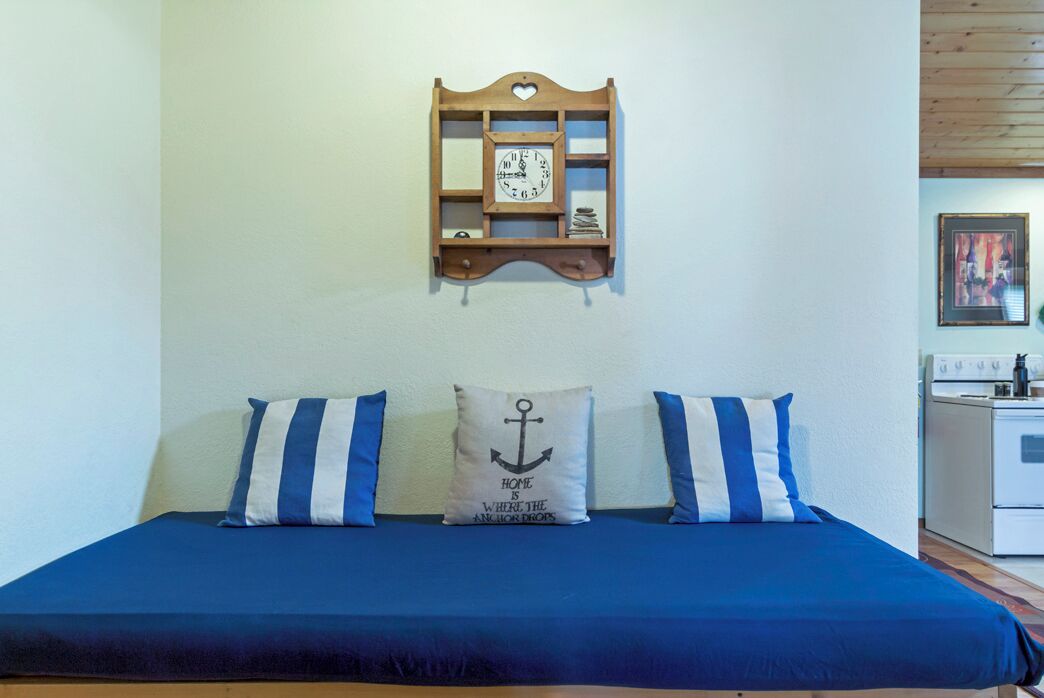 Een bed met blauw beddengoed, twee kussens met een anker dessin en een houten wandklok erboven.