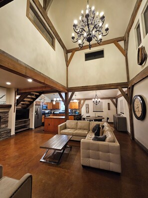 A spacious living area with a high ceiling, exposed wooden beams, a chandelier, a sofa, a coffee table, and a kitchen in the background. - Luxury Winter Ski Vacation Retreat in Big Sky - Hot Tub (Gallatin Gateway)