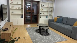 A modern living room with a grey sofa, a wooden coffee table, and a grey patterned rug. There are white shelves with decorative items and a dining area visible through a glass door. - Charming 2-bedroom apartment with WiFi, AC in fabulous Tiranë (Tiranë)