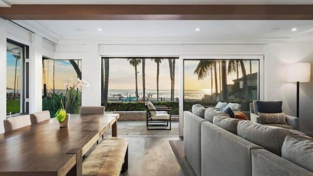 A modern living room with a large window overlooking a beach, a wooden dining table, and a grey sofa.