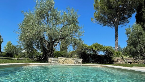 Uma piscina cristalina com uma fonte de pedra no centro, rodeada por árvores e um muro de pedra.