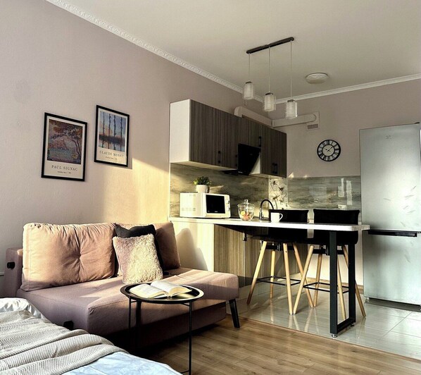 A modern living room with a sofa, a coffee table with a book, and a kitchen area with a microwave, a sink, and a refrigerator.
