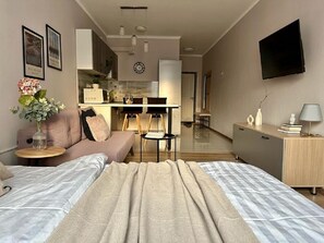 A modern hotel room with a large bed, a sofa, a small table with a vase of flowers, a desk with a lamp, and a TV mounted on the wall.