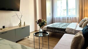 Une chambre d’hôtel moderne équipée d’un lit, d’un canapé, d’un téléviseur fixé au mur, d’une petite table avec un vase de fleurs et d’une fenêtre avec des rideaux.