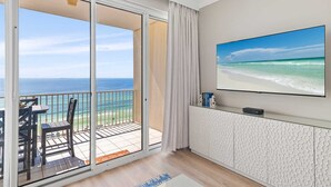 Ein Zimmer mit einer großen Schiebeglaswand, die auf einen Balkon mit Meerblick führt. An der Wand ist ein Flachbildfernseher angebracht und der Balkon ist mit einem kleinen Tisch und Stühlen ausgestattet.
