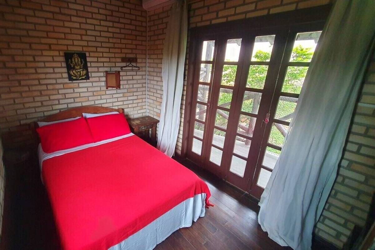 Une pièce avec un mur en briques, un lit recouvert d’une couverture rouge, une table de chevet en bois, un luminaire suspendu et une porte donnant sur un espace extérieur.