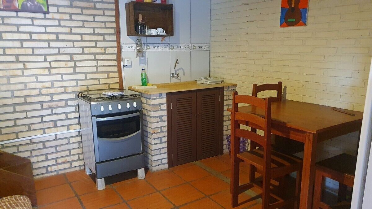 Uma cozinha compacta com fogão a gás, forno e uma pia pequena. Há uma cadeira de madeira e uma mesa, e uma prateleira acima da pia.