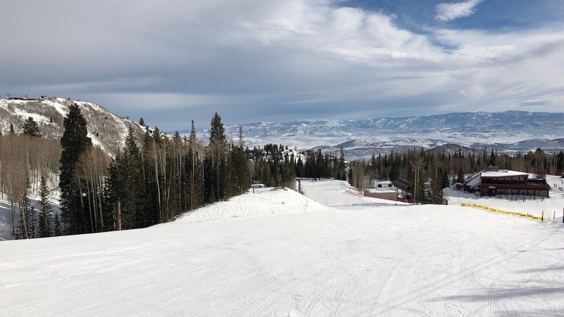 Pista de esquí cubierta de nieve con una cabaña y montañas al fondo.