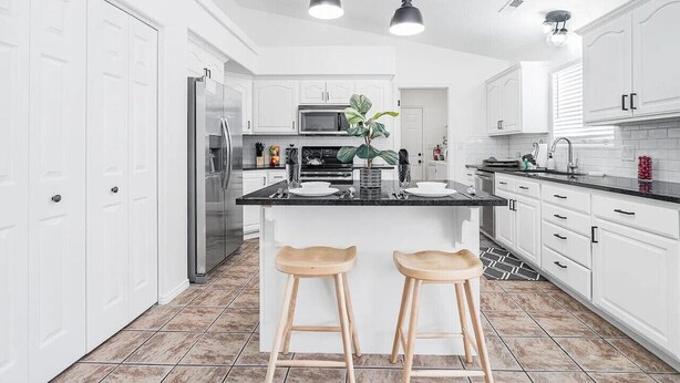 A modern kitchen with white cabinetry, a stainless steel refrigerator, a black countertop, two wooden stools, and pendant lighting. - Massive Backyard with Private Pool and Spa (Hurricane)