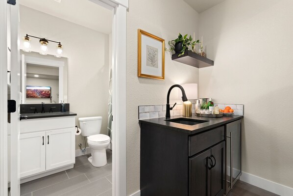 Une salle de bain moderne avec des toilettes blanches, un lavabo noir doté d’un robinet doré et un miroir avec éclairage intégré. On y trouve une étagère avec des plantes et un tableau encadré accroché au mur.