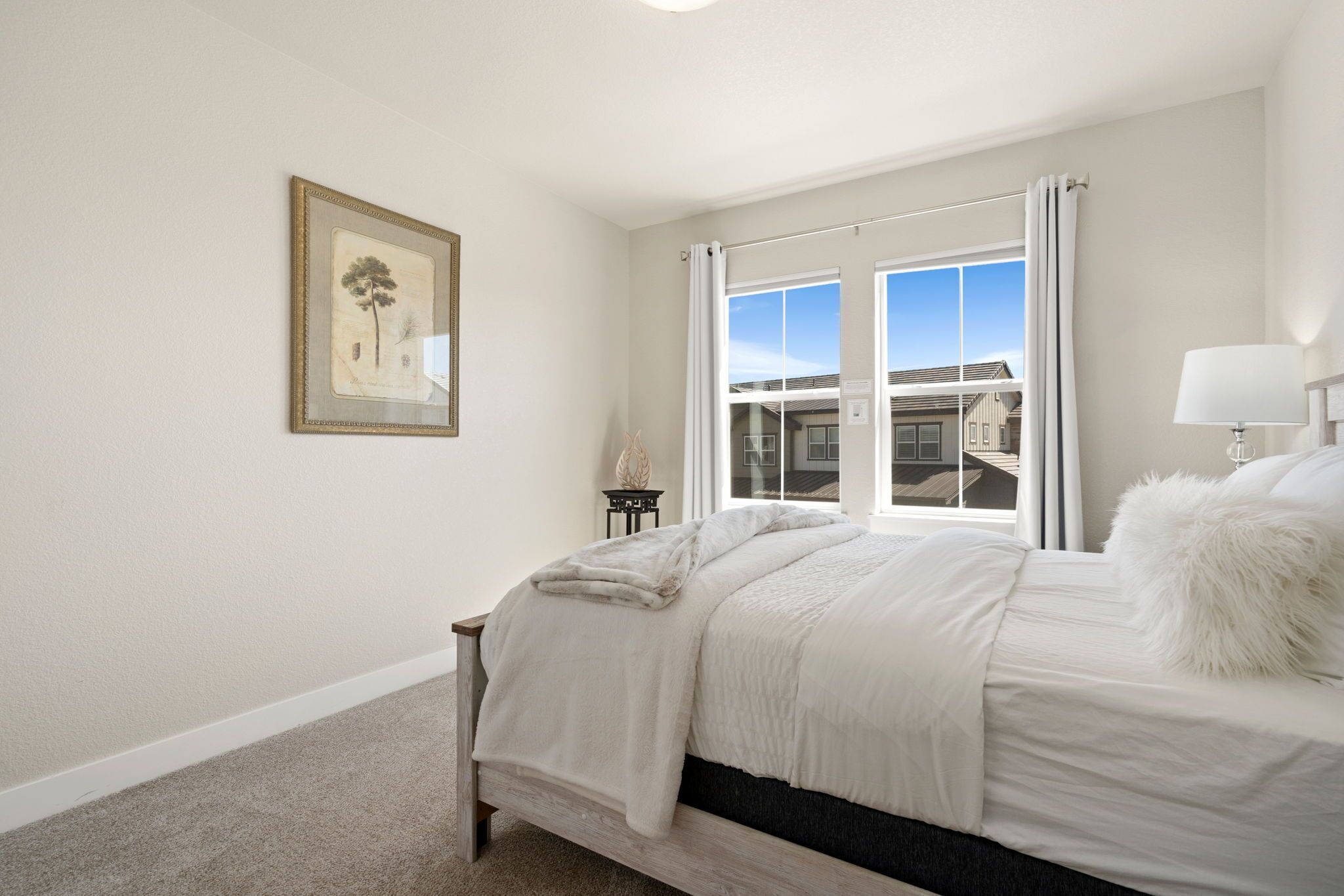 A bedroom with a large bed, a window with curtains, a small table with a lamp, and a framed picture on the wall.