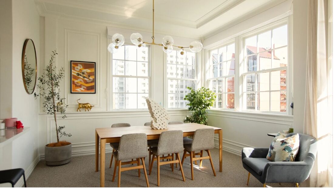 Un comedor con una mesa de madera, cuatro sillas, una araña y ventanas amplias con vistas a edificios.