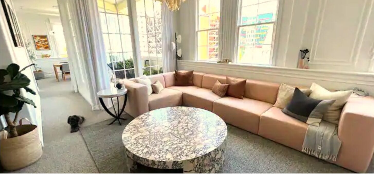Een ruime woonkamer met een grote beige modulaire bank, een ronde marmeren salontafel en een klein bijzettafeltje.