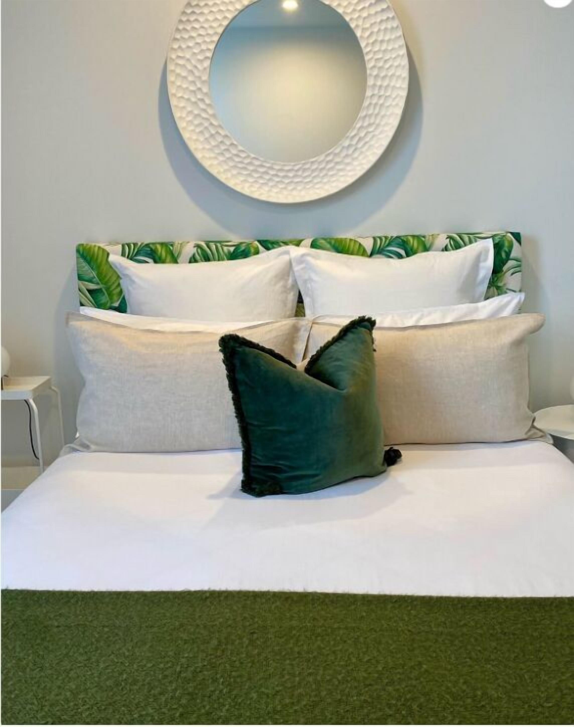 A bed with white bedding and a green pillow, a round mirror on the wall, and a green wall.