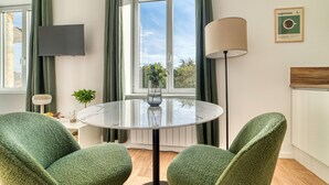 Ein modernes Wohnzimmer mit einem runden Glastisch, zwei grünen Polstersesseln, einem flachbildfernseher an der Wand, einer Stehlampe und einem Fenster mit Vorhängen.