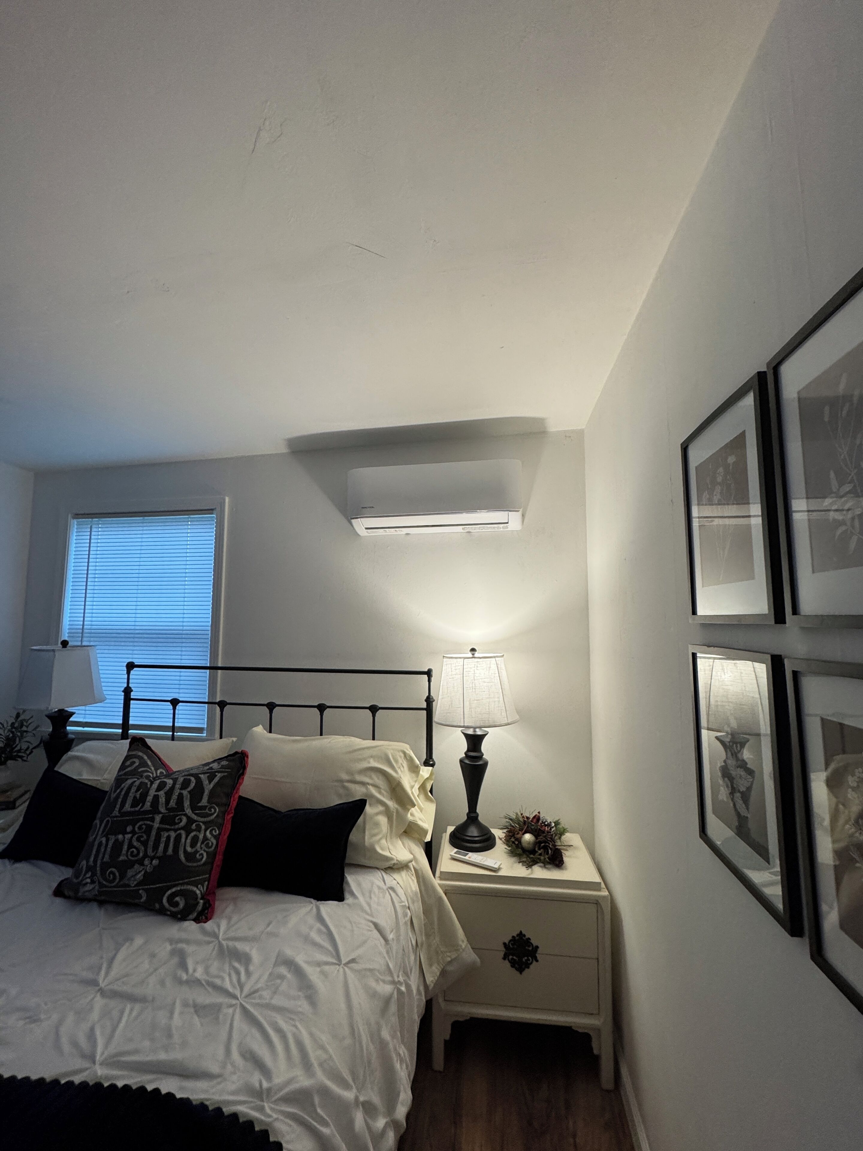 Une chambre à coucher avec un lit, une table de chevet, une lampe, une fenêtre avec des stores et des tableaux encadrés accrochés au mur.