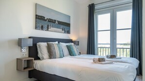 A double bed with white linens, two bedside tables with lamps, a painting of a coastal scene, and a large window with curtains.
