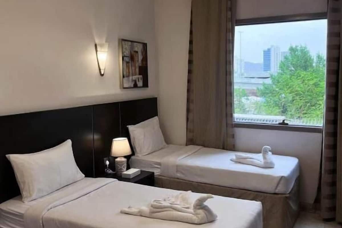 A hotel room with two beds, a nightstand with a lamp, a painting on the wall, and a window with a view of buildings and trees.