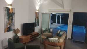 Ein Wohnzimmer mit Flachbildfernseher, Couchtisch aus Holz und einer Couch mit Kissen. An der Wand hängt ein großes Gemälde und durch Glastüren ist ein Swimmingpool zu sehen.