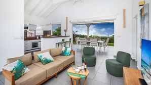 Ein modernes Wohnzimmer mit einer Couch, Sesseln und einem Essbereich. Im Hintergrund ist eine Küche zu sehen, und ein großes Fenster bietet einen Blick ins Freie.