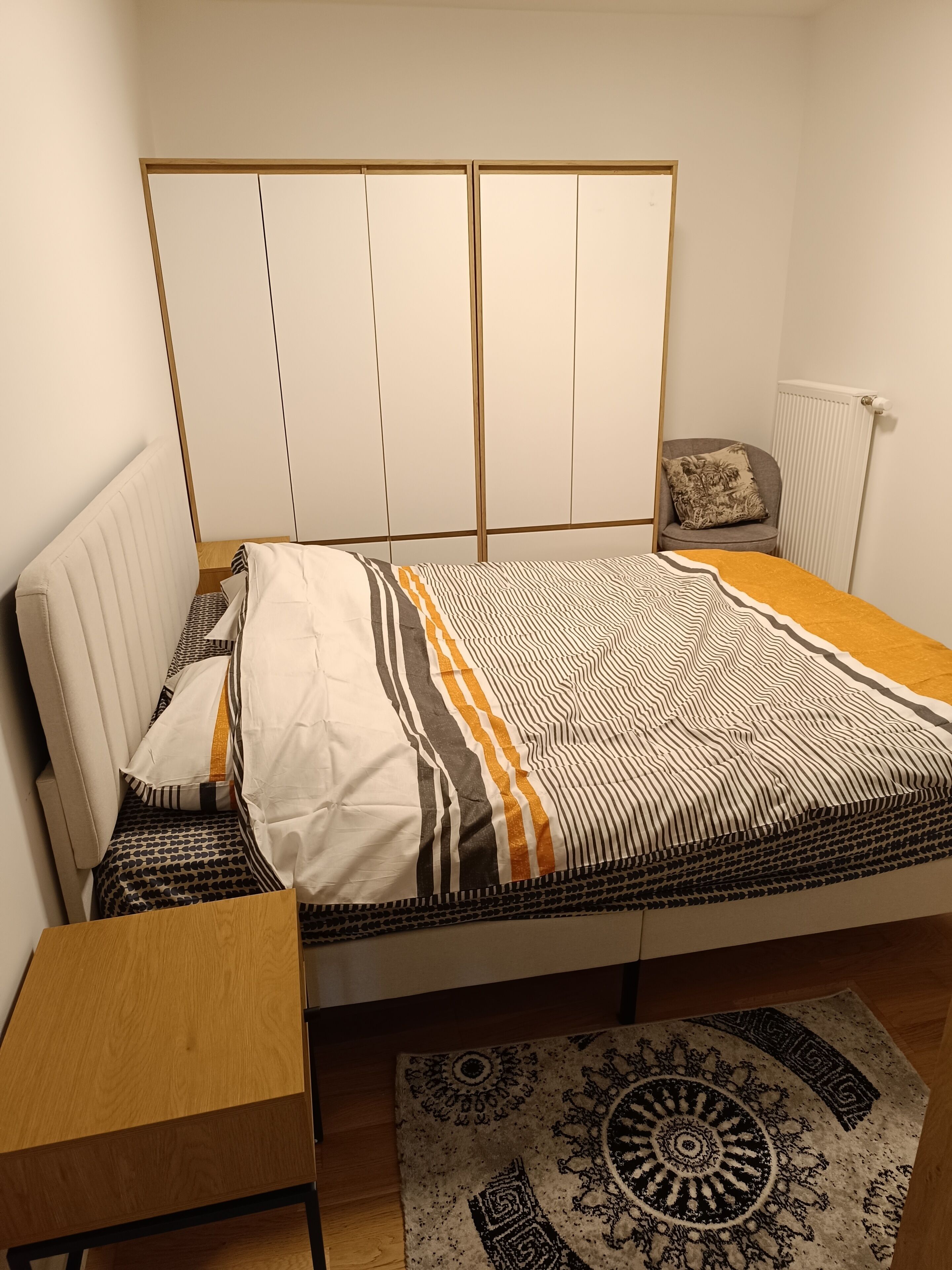 Ein Schlafzimmer mit einem Bett, einem Holznachttisch, einem Stuhl, einem gemusterten Teppich und einer Heizung.