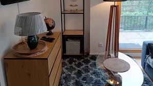 Moderni olohuone, jossa on puinen kaappi, televisio, koriste-esineillä täytetty hylly, lamppu ja näkymä puita kohti ikkunasta.
