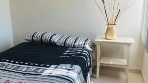 Un lit bien fait, recouvert d’une couvre-lit à rayures et agrémenté d’un oreiller, une petite table de chevet blanche sur laquelle est posé un vase contenant des plantes séchées, et un mur sobre.