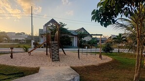Um parquinho com balanços e estrutura de escalada, rodeado por um espaço pavimentado e palmeiras.