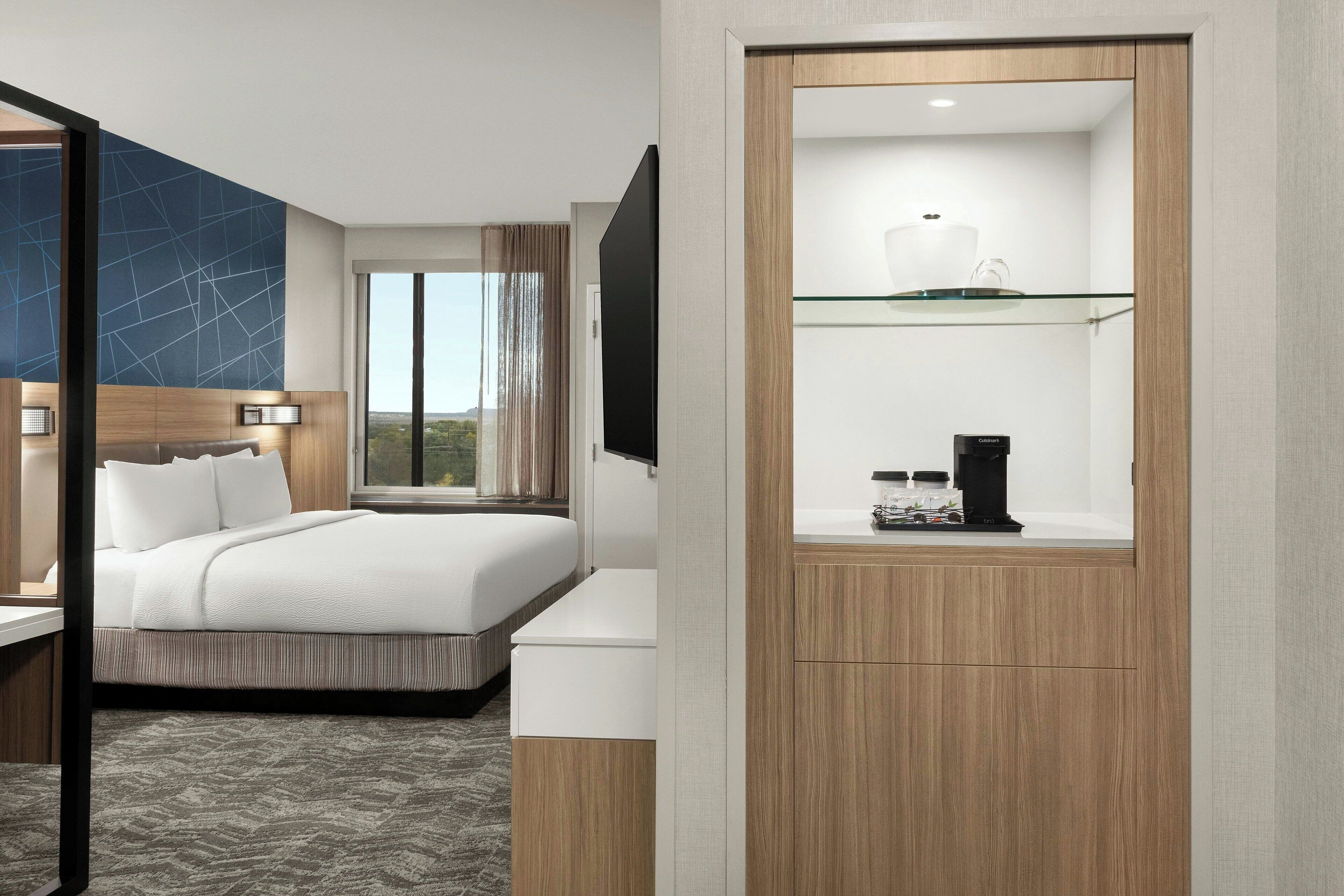 A modern hotel room with a large bed, a desk, a coffee machine, and a view of the outdoors.