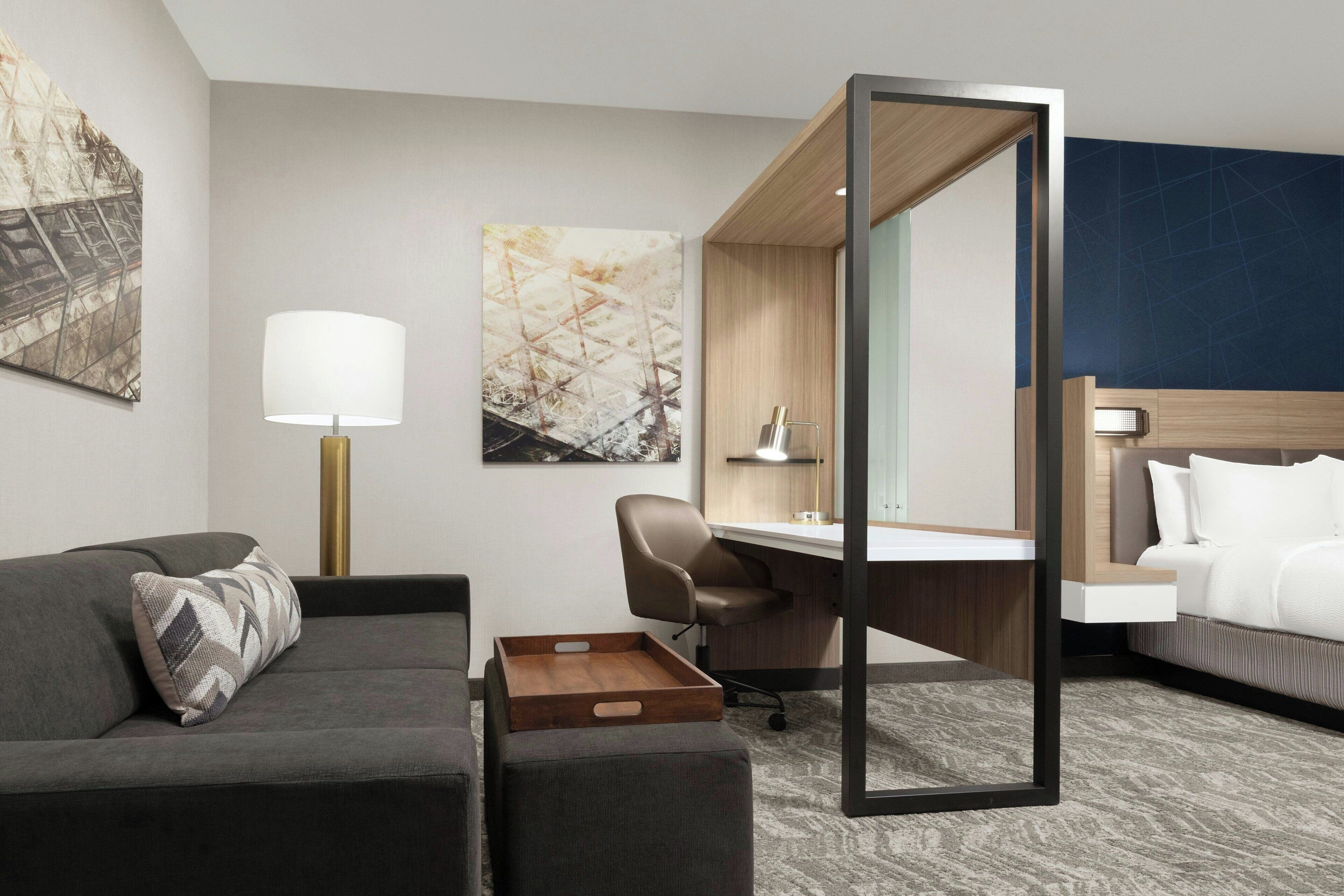 A modern hotel room with a grey sofa, a wooden desk, a white bed, and a large mirror.