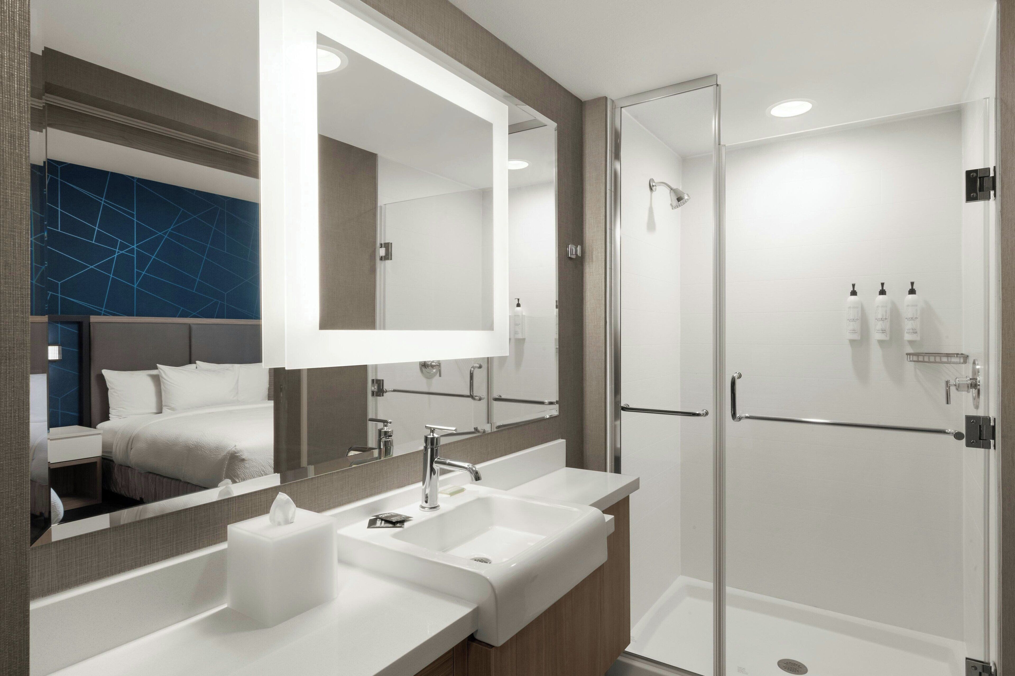 A modern bathroom with a large mirror, two sinks, a shower, and a bedside table with a lamp.