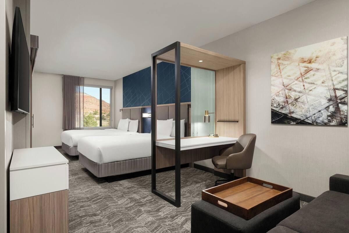 Ein modernes Hotelzimmer mit zwei Betten, einem Schreibtisch, einem Stuhl und einem großen Fenster mit Blick auf eine gebirgige Landschaft.