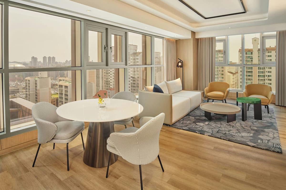 A modern hotel room with a large window, a round dining table, two chairs, a sofa, and a small coffee table.