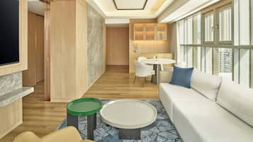 A modern hotel room with a large TV, a sofa, a dining area, and a kitchen.