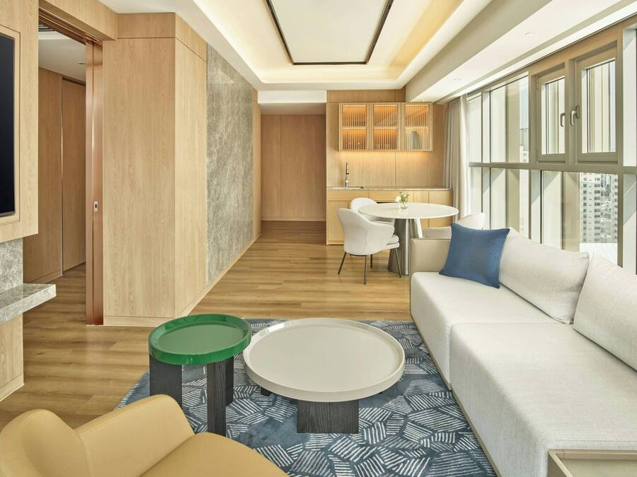 A modern hotel room with a large TV, a sofa, a dining area, and a kitchen.