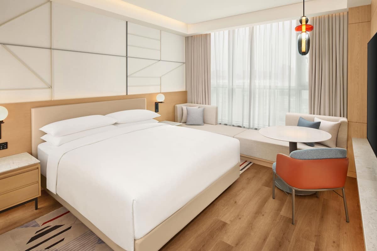 A modern hotel room with a large bed, a seating area with a table and chairs, and a large window with curtains.