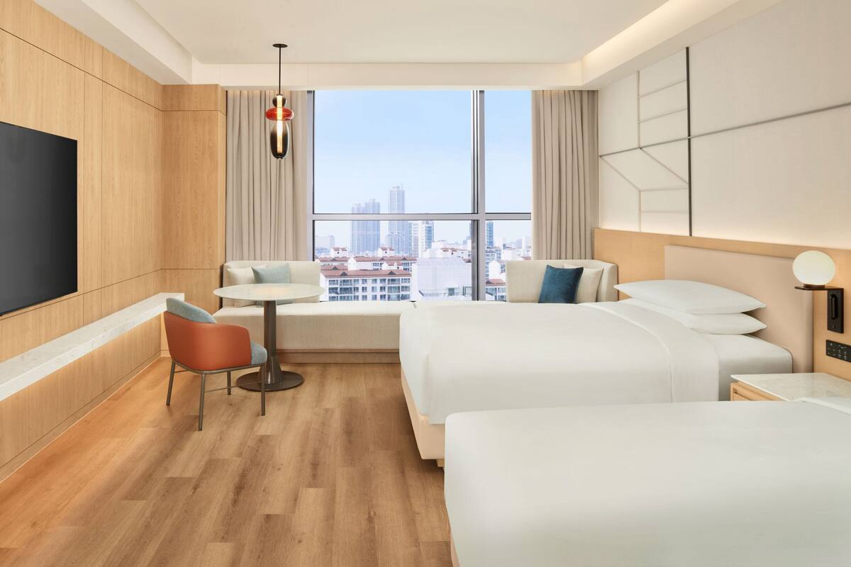 A modern hotel room with two beds, a TV, a dining table, and a city view.