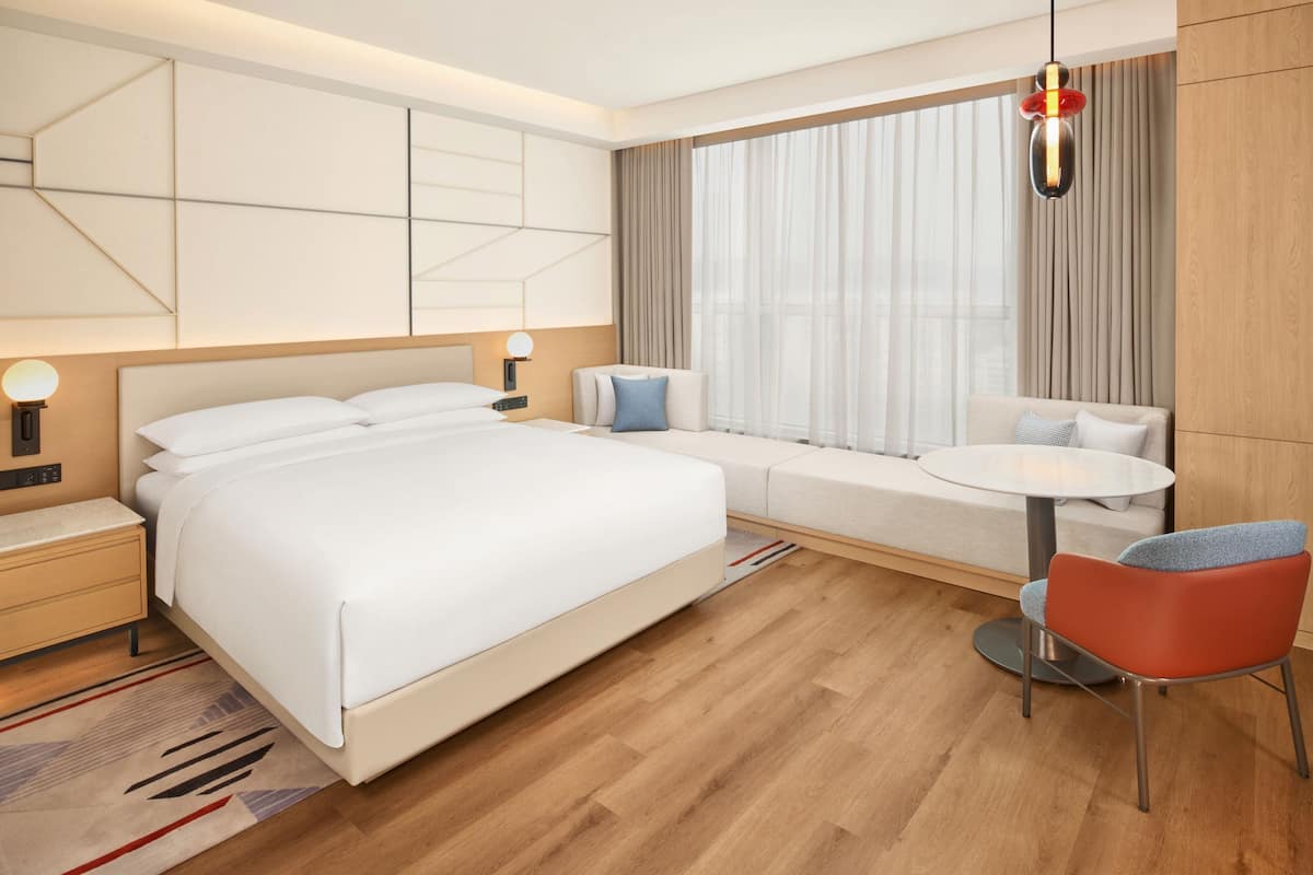 A modern hotel room with a large bed, a bedside table, a small round table, a chair, and a window with curtains.