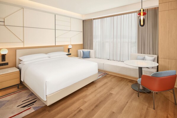 Ein modernes Hotelzimmer mit einem großen Bett, einem Nachttisch, einem kleinen runden Tisch, einem Sessel und einem Fenster mit Vorhängen.