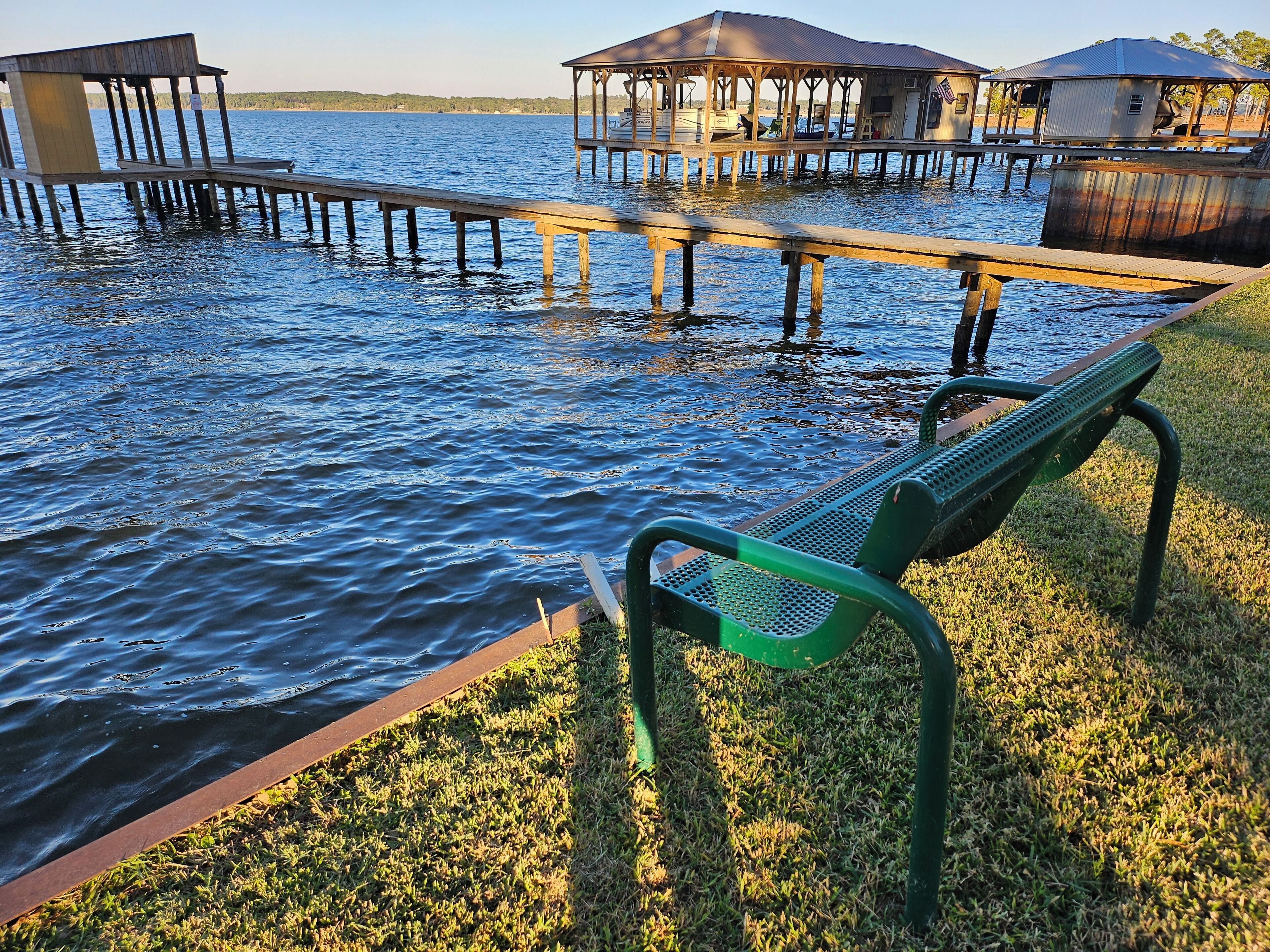 A wooden dock extending into a body of water with a grassy area and a metal bench in the foreground.