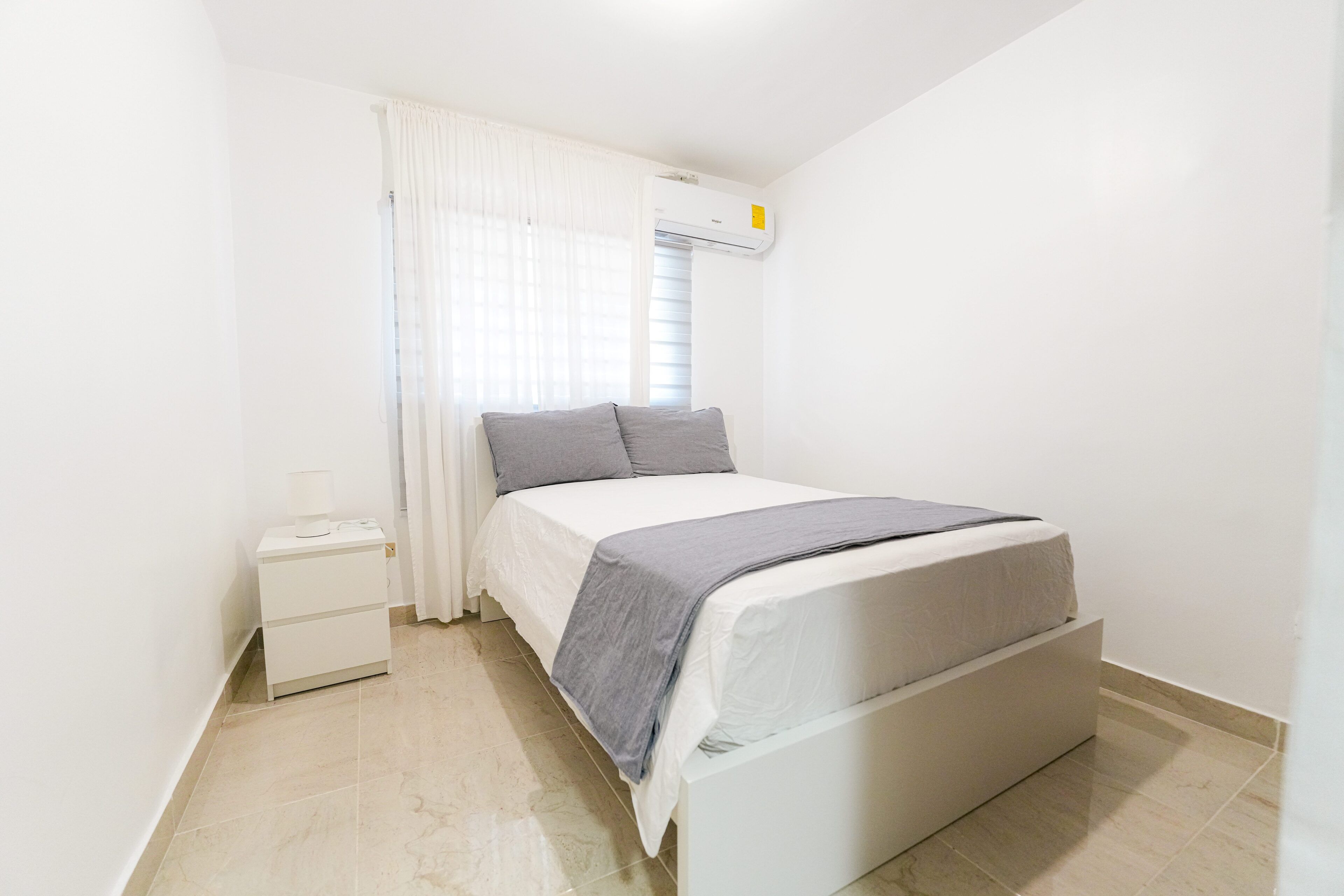 A bedroom with a bed, a nightstand, a window with curtains, and an air conditioning unit.