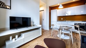 Un salon moderne doté d’un téléviseur à écran plat, d’un meuble de rangement blanc, d’un coin repas avec une table ronde et des chaises, et d’une cuisine avec des armoires blanches et un plan de travail en bois.