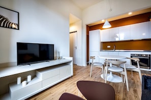 A modern living room with a flat-screen TV, a white entertainment unit, a dining area with a round table and chairs, and a kitchen with white cabinets and a wooden countertop.