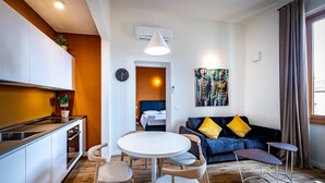 Une cuisine moderne avec des meubles blancs, un coin repas avec une table ronde et des chaises, un canapé bleu avec des coussins jaunes et un tableau au mur.