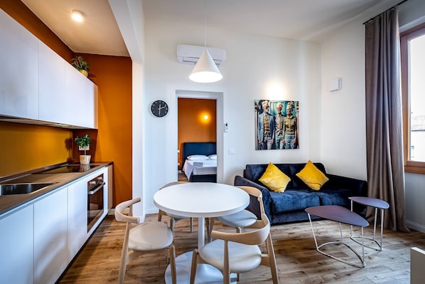 A modern kitchen with white cabinets, a dining area with a round table and chairs, a blue sofa with yellow cushions, and a painting on the wall.