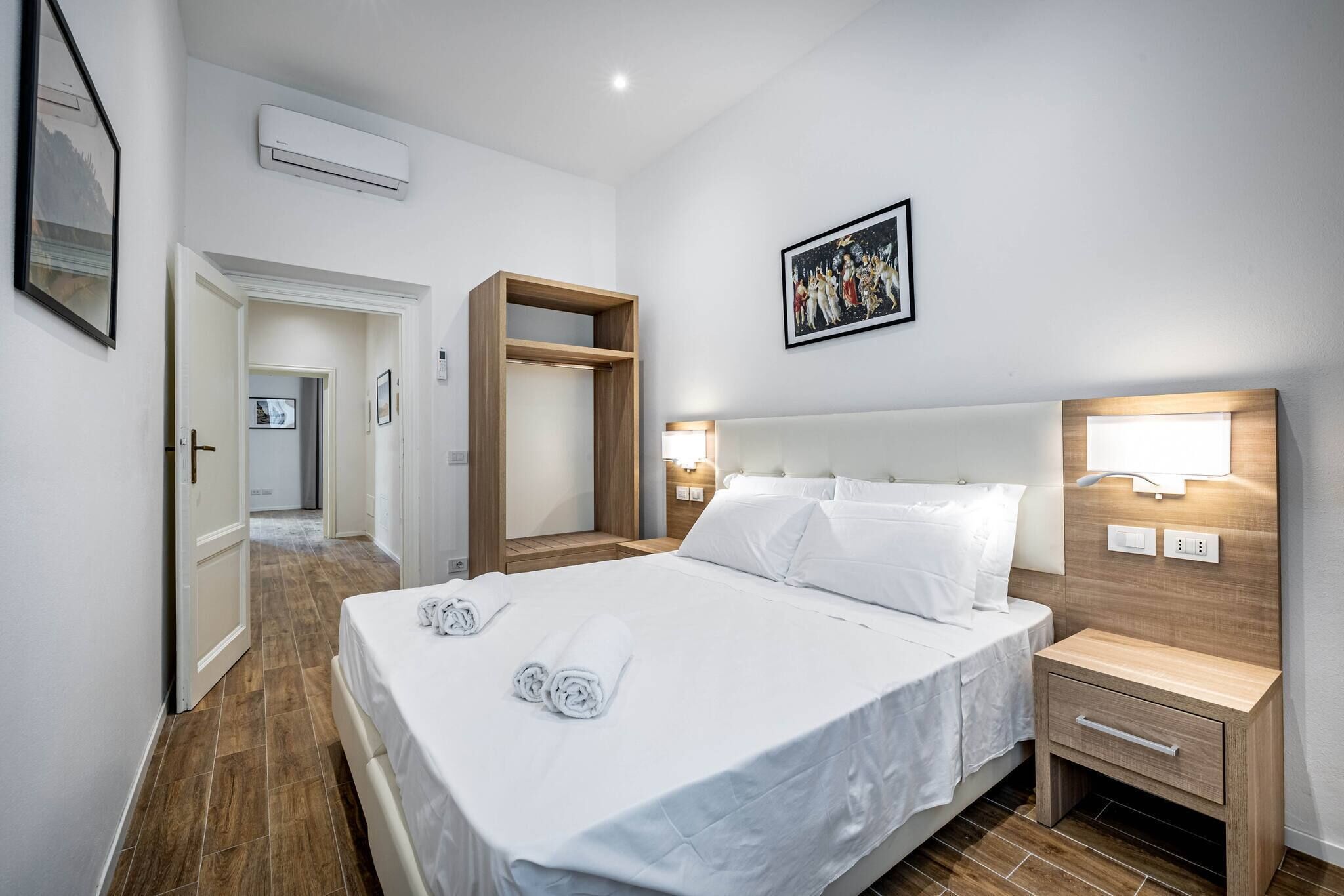 Hotelkamer met een groot bed, een houten nachtkastje, een ingebouwde airconditioner en een lijst met een afbeelding boven het bed.
